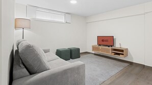 Ein modernes Wohnzimmer mit einem grauen Sofa, einem kleinen grünen Hocker, einem Holz-TV-Möbel mit einem Flachbildfernseher, der „GUEST READY“ anzeigt, und einer Stehlampe.