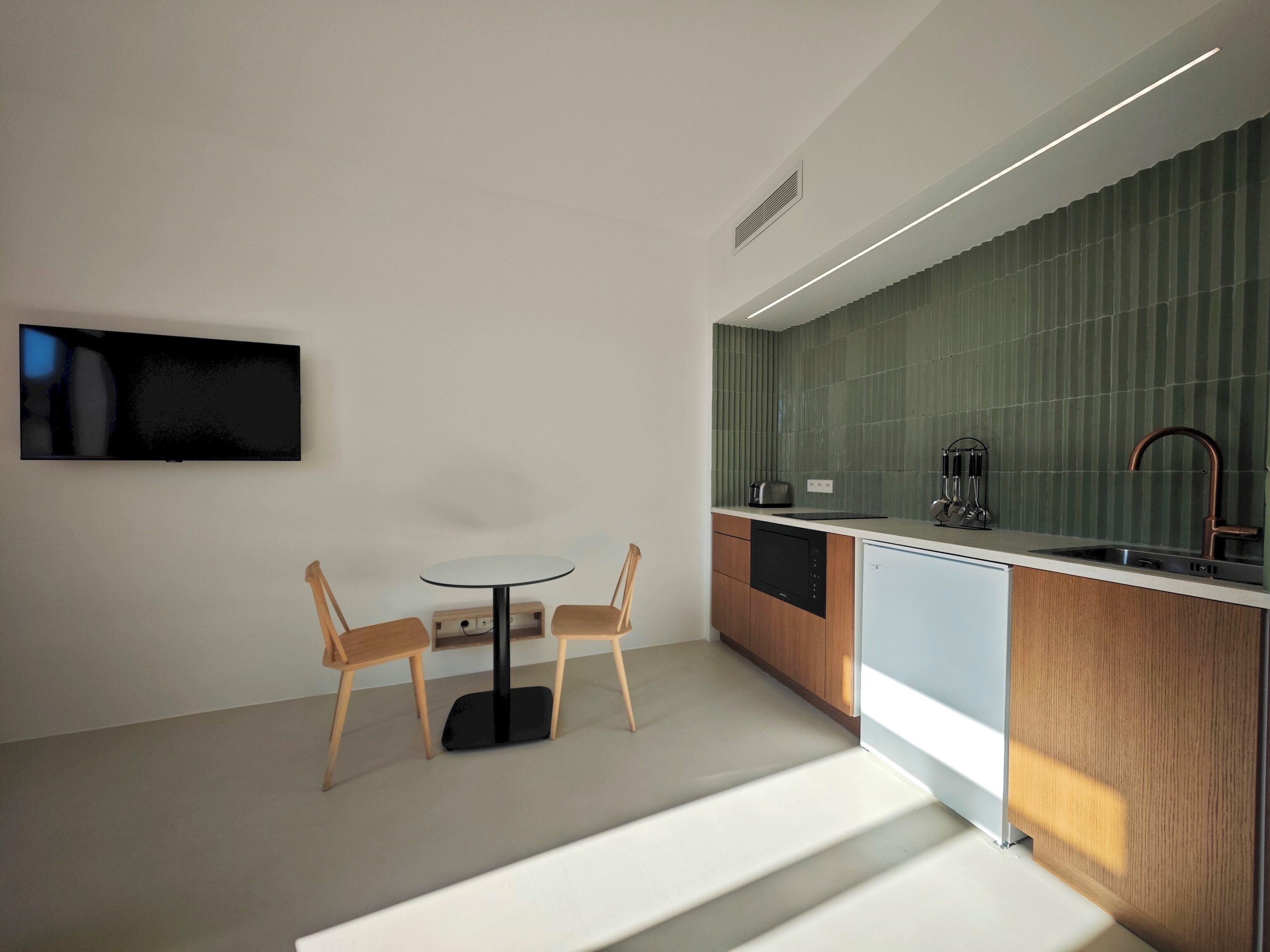 A modern kitchen with a dining area, featuring a flat-screen TV, wooden chairs, a round table, and a green backsplash.