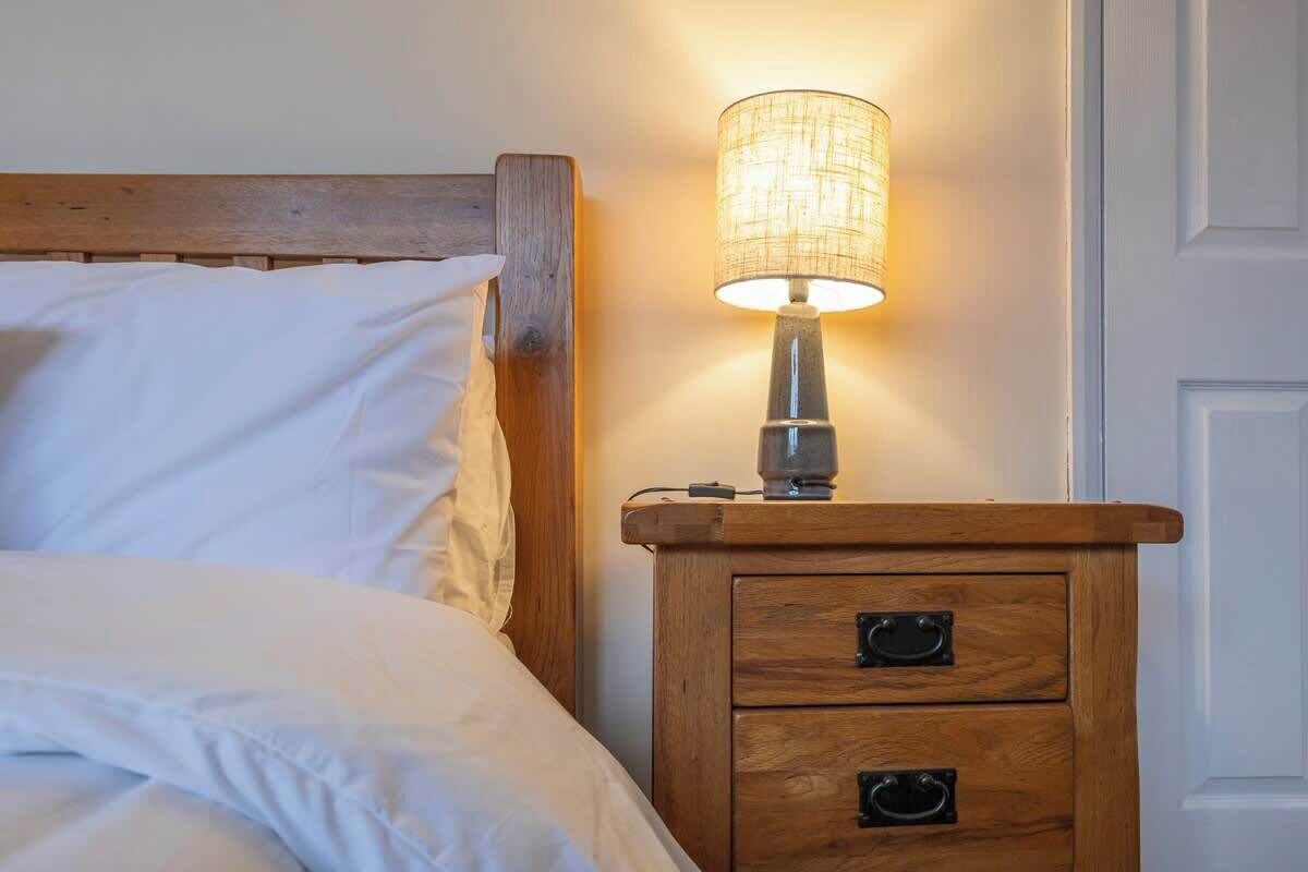 A neatly made bed with white linens, a wooden nightstand with a lamp, and a white door in the background.