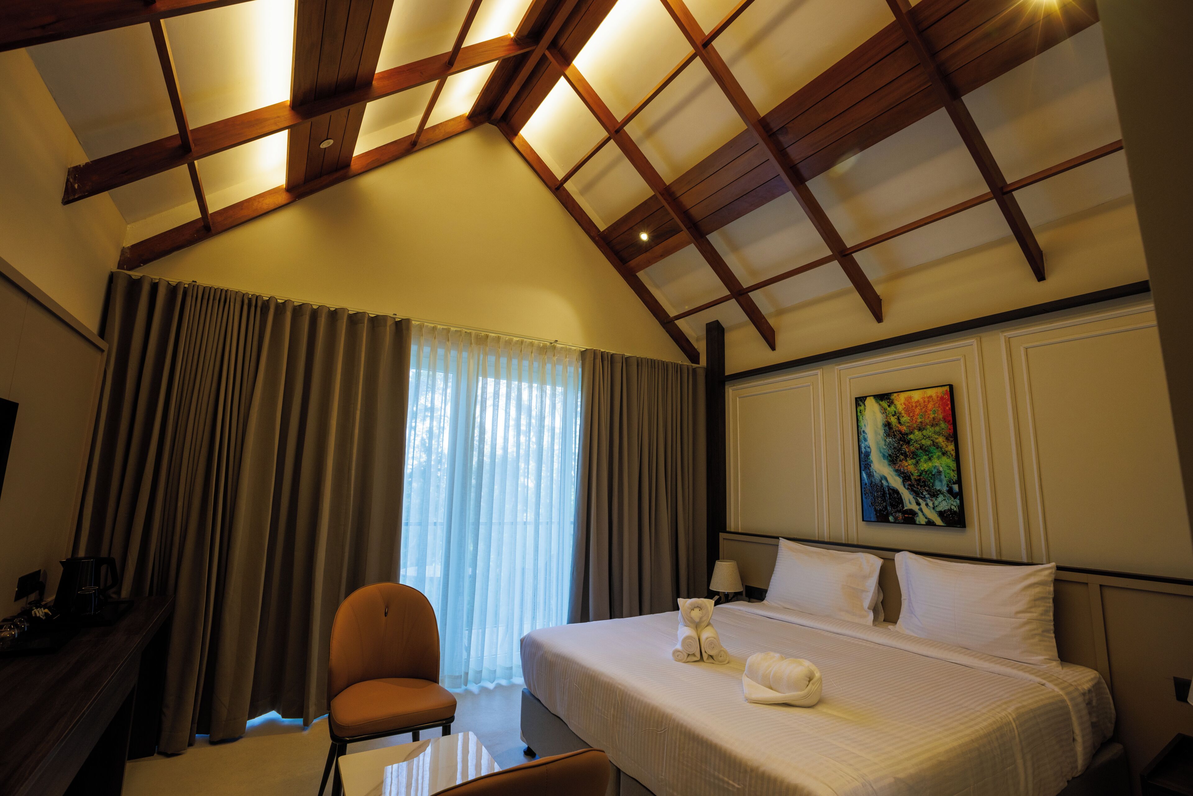 Habitación de hotel con techo alto, cama con ropa blanca, una silla, escritorio con lámpara y un cuadro en la pared.