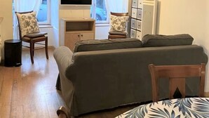 A cozy living room with a sofa set, a dining table with a floral tablecloth, and a flat-screen TV mounted on the wall.
