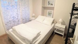 A bedroom with a bed, two bedside tables, a nightstand, a wall-mounted shelf with pictures, and a curtain.