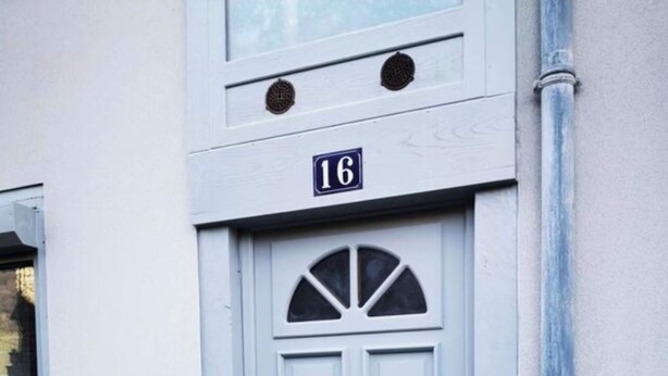 Une porte bleu clair, surmontée du numéro « 16 », est intégrée à un immeuble dont l’entrée est pavée de pierres.