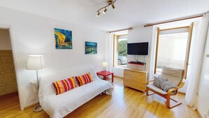 Une pièce avec un canapé blanc, un coussin à rayures, une chaise en bois, un téléviseur posé sur une étagère et des œuvres d’art accrochées au mur.