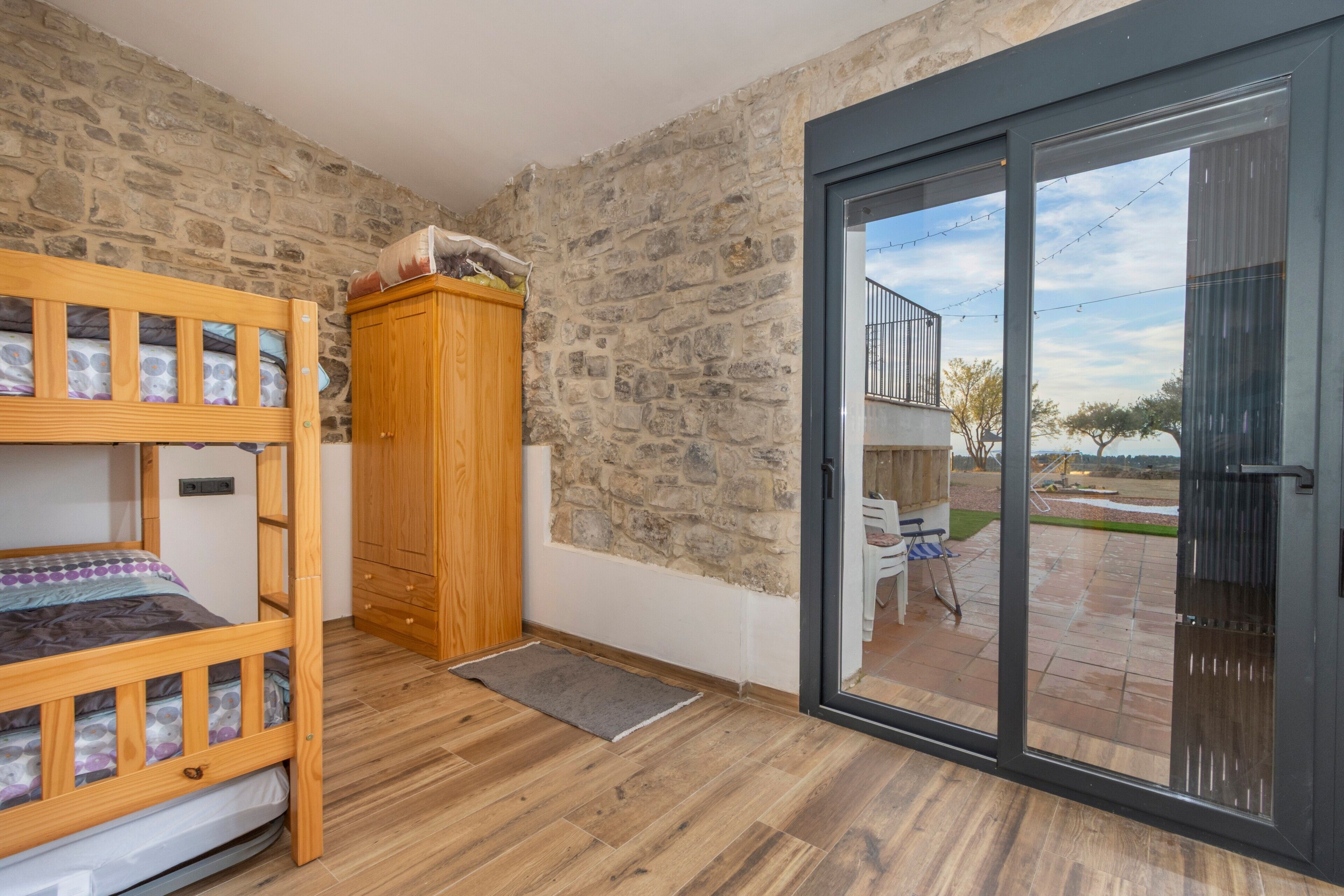 Una habitación con literas, un armario de madera, pared de piedra y una puerta corrediza de vidrio que da a un espacio exterior.