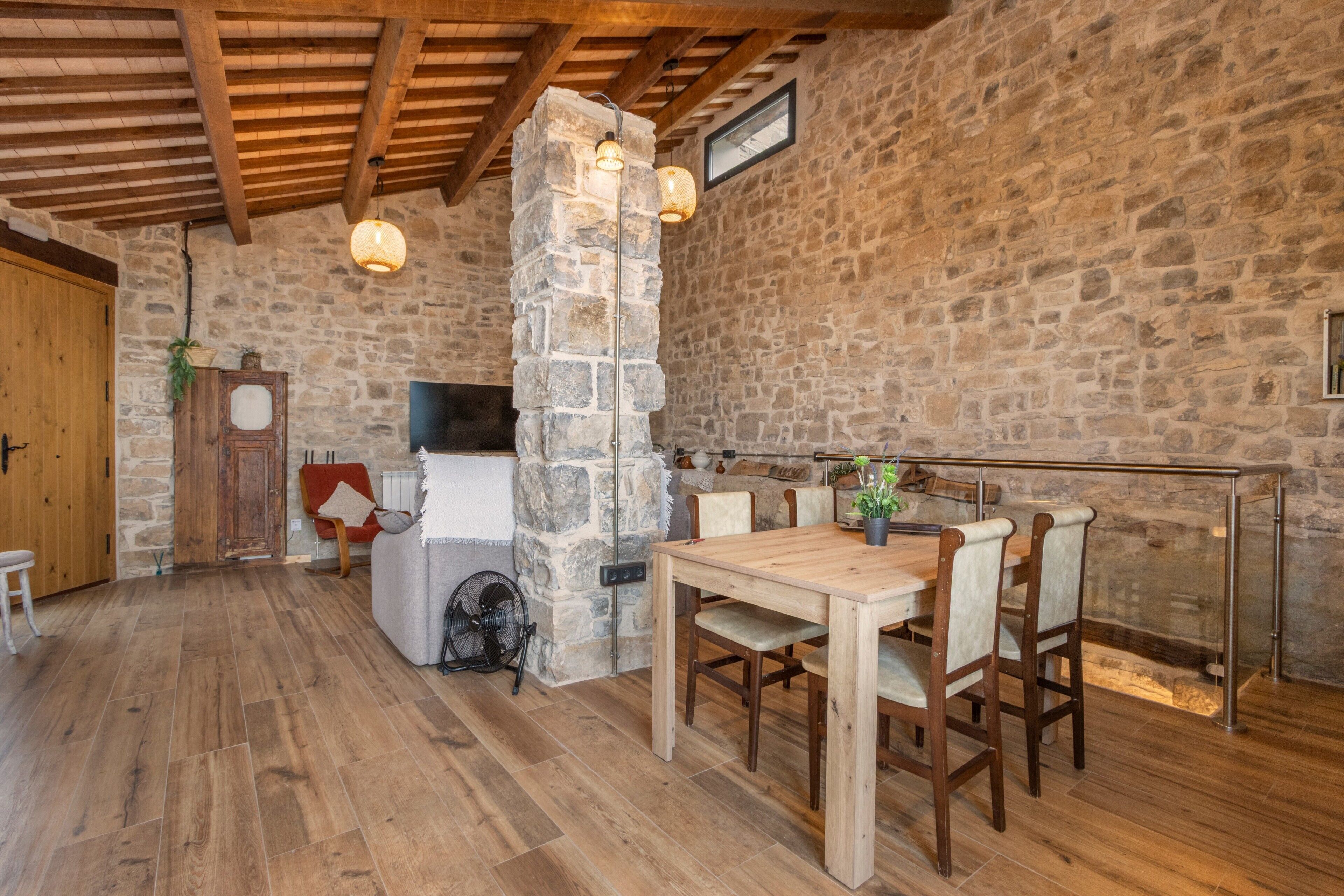 Un interior rústico con pared de piedra, vigas de madera en el techo, un pilar de piedra, una mesa de comedor de madera y sillas.