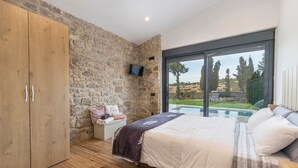 Een slaapkamer met een stenen muur, een houten garderobe, een bed met witte beddengoed, een televisie en uitzicht op een zwembad.