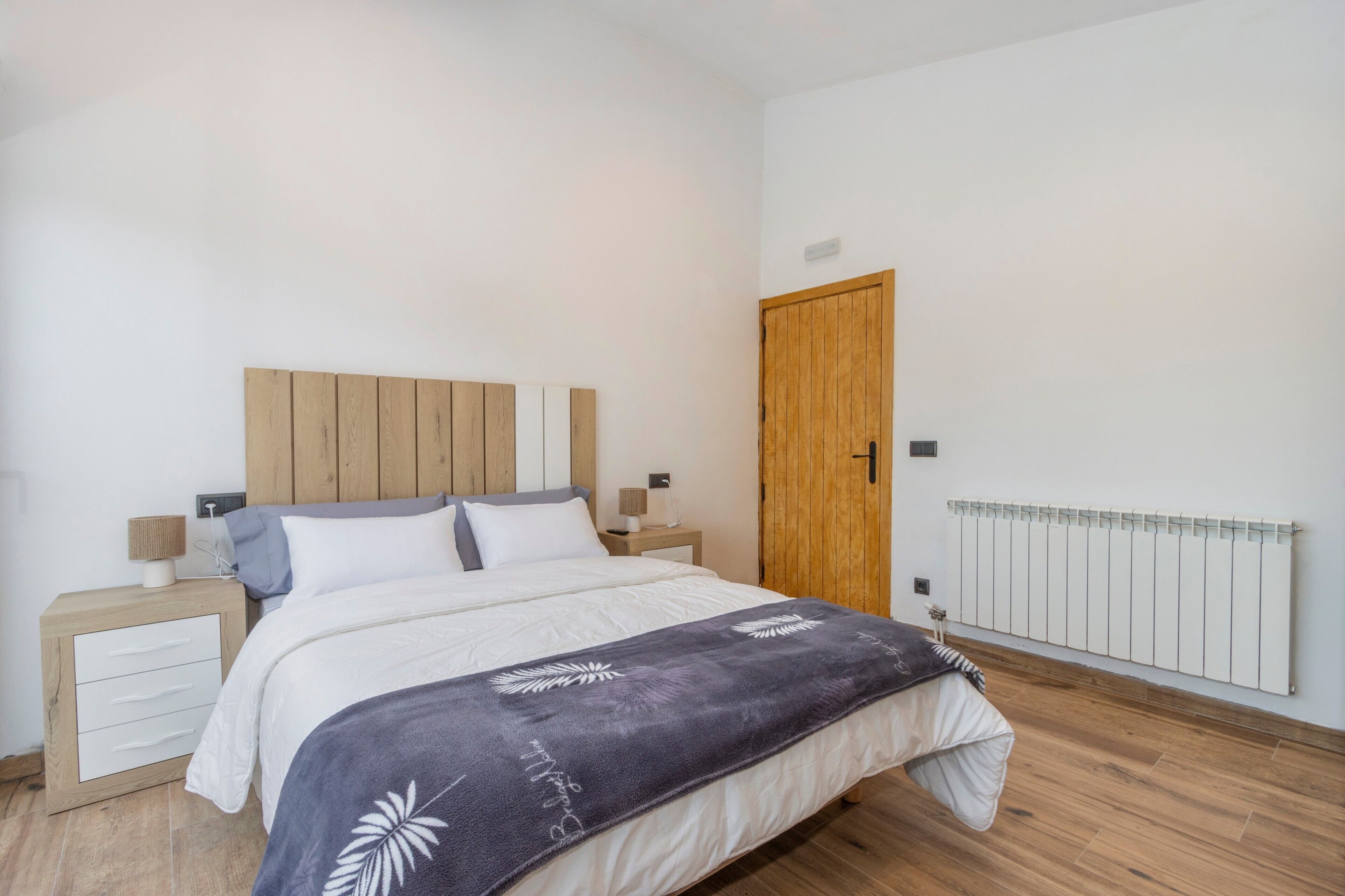 A bedroom with a wooden headboard, a bed with white bedding, a nightstand with a lamp, a wooden door, and a radiator.