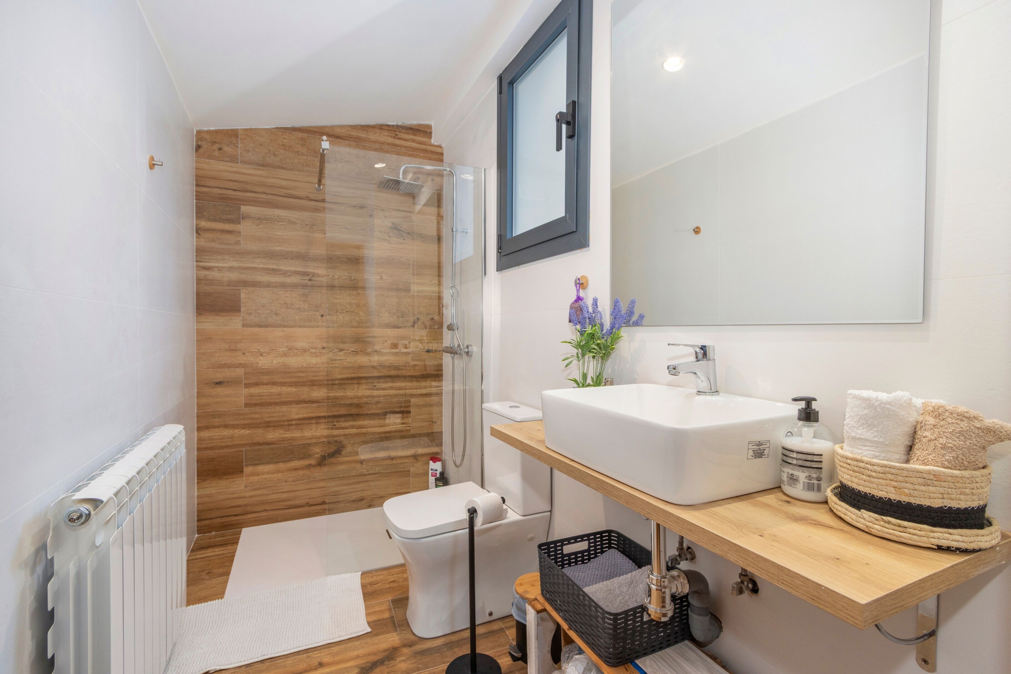 Un baño moderno con cabina de ducha de madera, un lavabo blanco, un espejo y un estante con toallas y artículos de aseo.