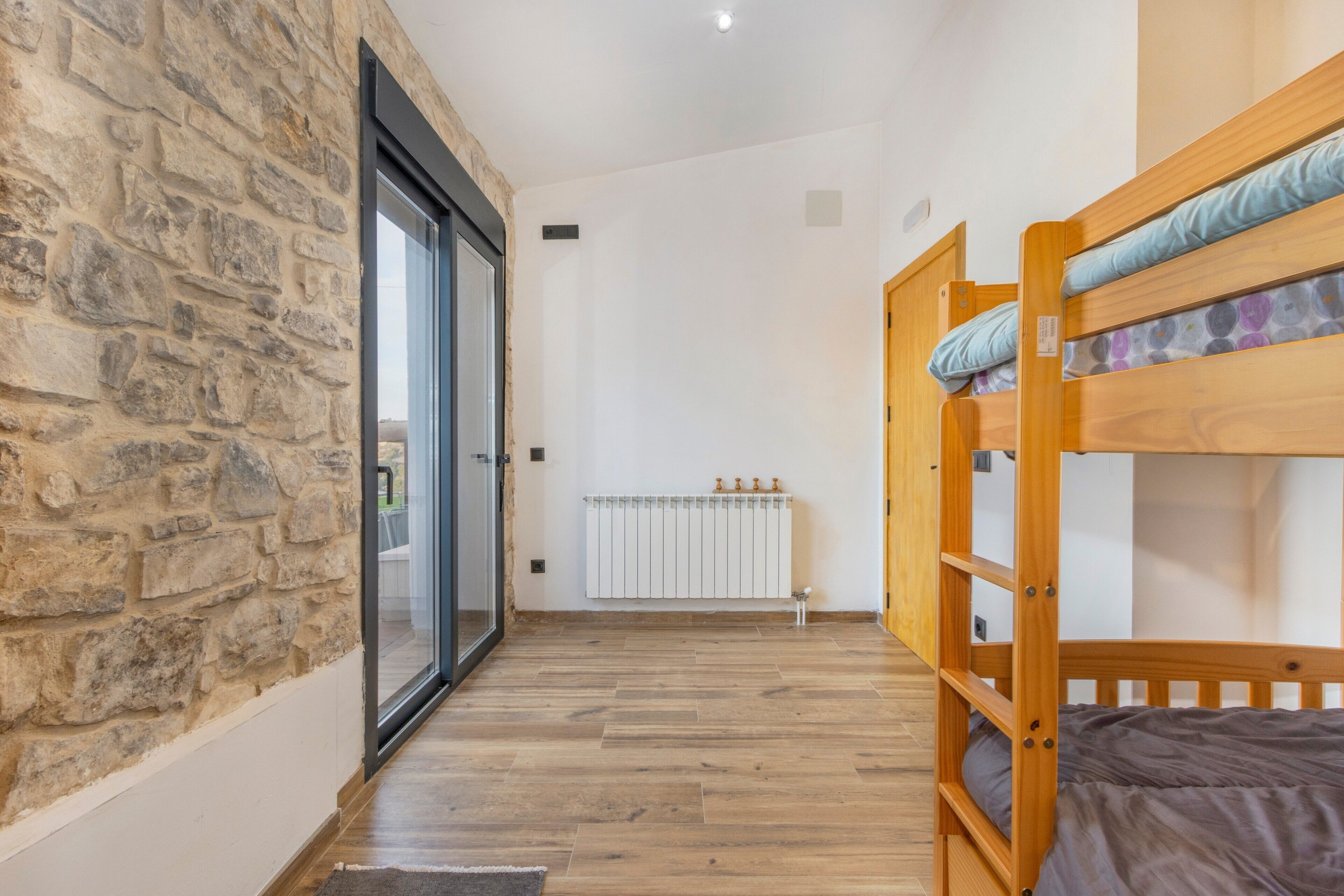 A room with a stone wall, wooden flooring, a bunk bed, and a sliding glass door leading to a balcony.