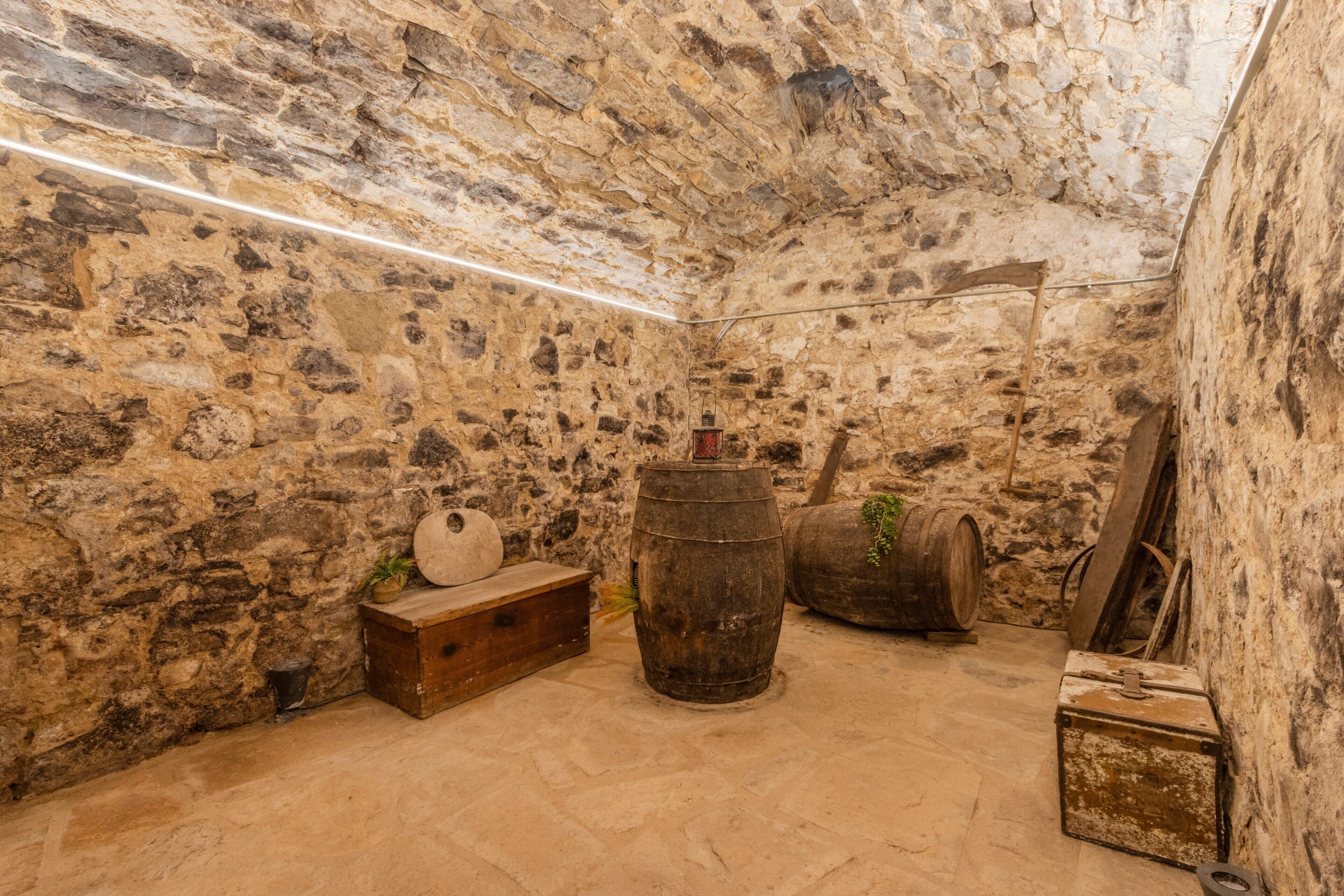 Una habitación con paredes de piedra, un banco de madera, dos barriles y una caja de herramientas.