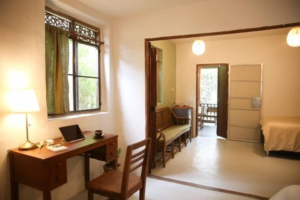 Une pièce comprenant un bureau, une chaise, un lit et donnant sur l’extérieur par une porte ouverte.