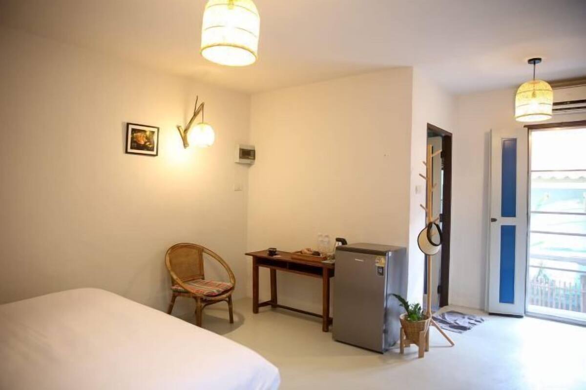 Une chambre d’hôtel comprenant un lit, un bureau, une chaise, un réfrigérateur, un luminaire fixé au mur et une porte donnant sur un espace extérieur.