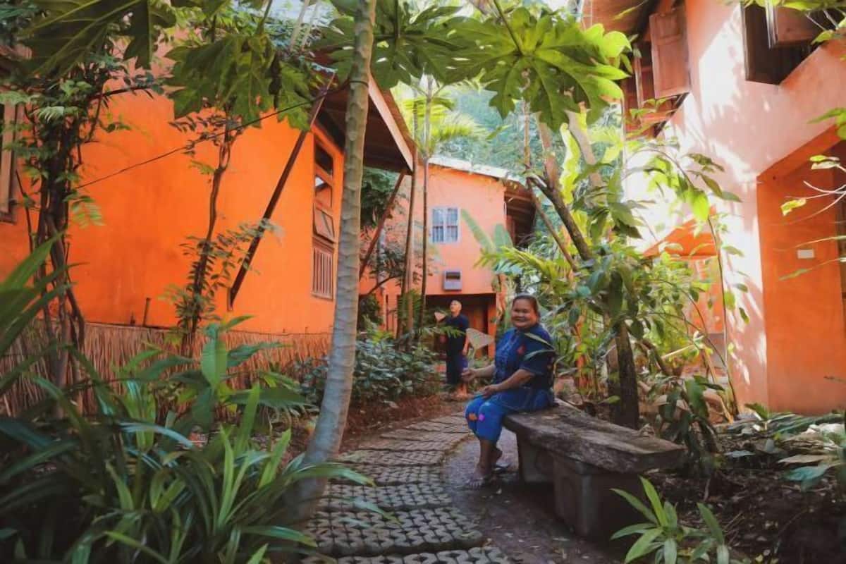 Un étroit sentier mène à un bâtiment aux murs orange, entouré d’une végétation luxuriante et bordé d’un chemin pavé de pierres.