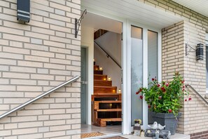 Une façade en briques, une porte d’entrée en verre, un escalier en bois et un pot de fleurs avec des fleurs rouges.