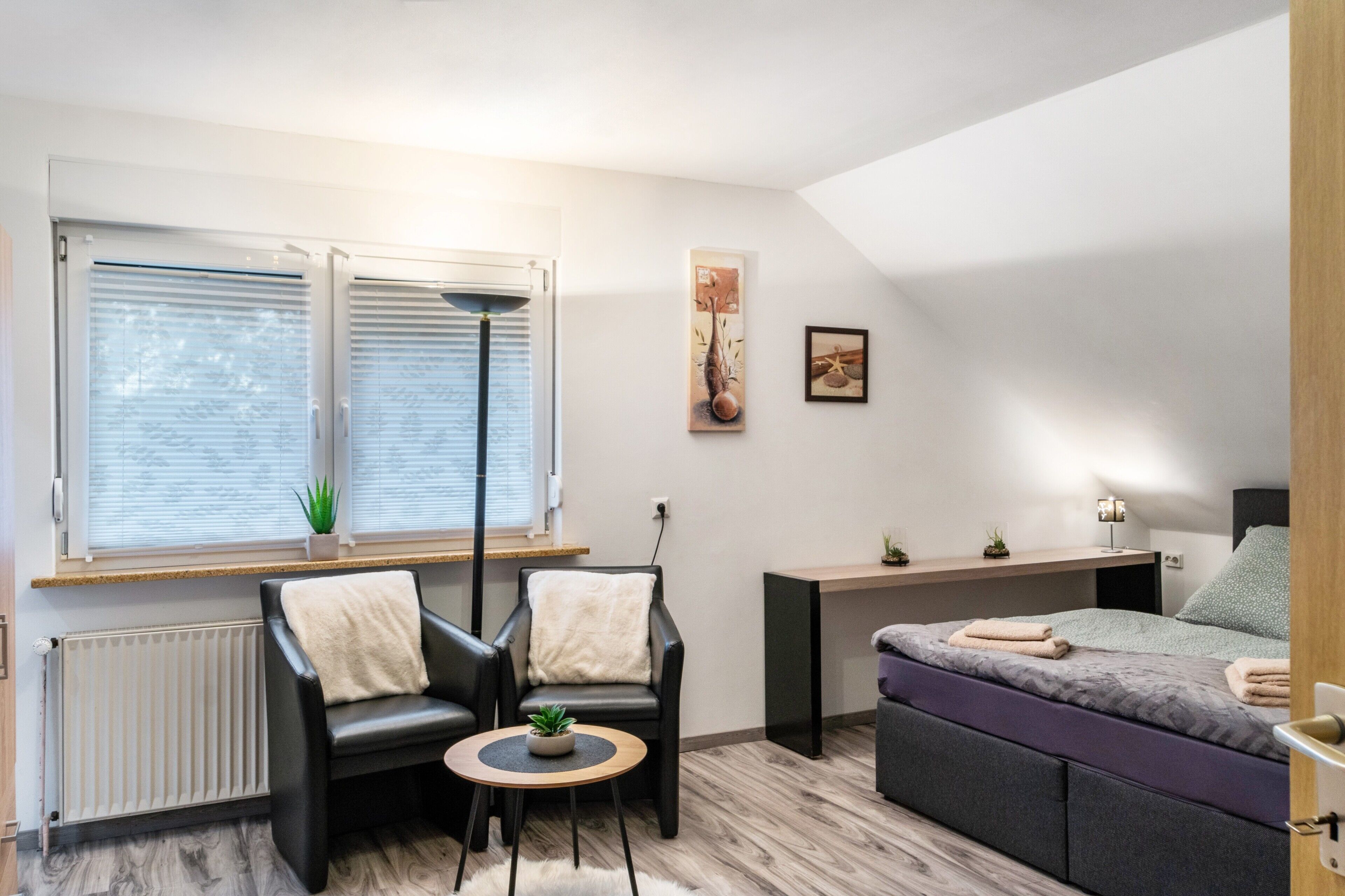 A modern room with a bed, two chairs, a small table, a radiator, a window with blinds, and a wall-mounted shelf.