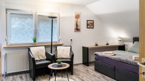 Een moderne kamer met een bed, twee stoelen, een tafeltje, een radiator, een raam met jaloezieën en een wandplank.