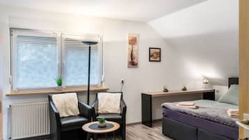 A modern room with a bed, two chairs, a small table, a radiator, a window with blinds, and a wall-mounted shelf.