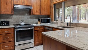 A kitchen with wooden cabinets, a stainless steel oven, a microwave, and a large window overlooking a forest. - The Meadow House in Sunriver (Sunriver)