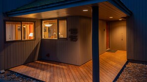 A covered outdoor area with wooden flooring, a small seating area, and a door leading to a building with a number on the wall. - The Meadow House in Sunriver (Sunriver)