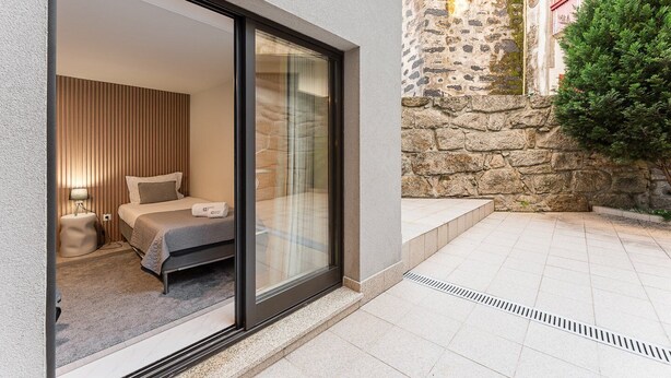 A modern hotel room with a large sliding glass door leading to a balcony. The room features a bed with a grey bedspread, a bedside table with a lamp, and a window with curtains. - GuestReady - Charming Porto's nook (Porto)