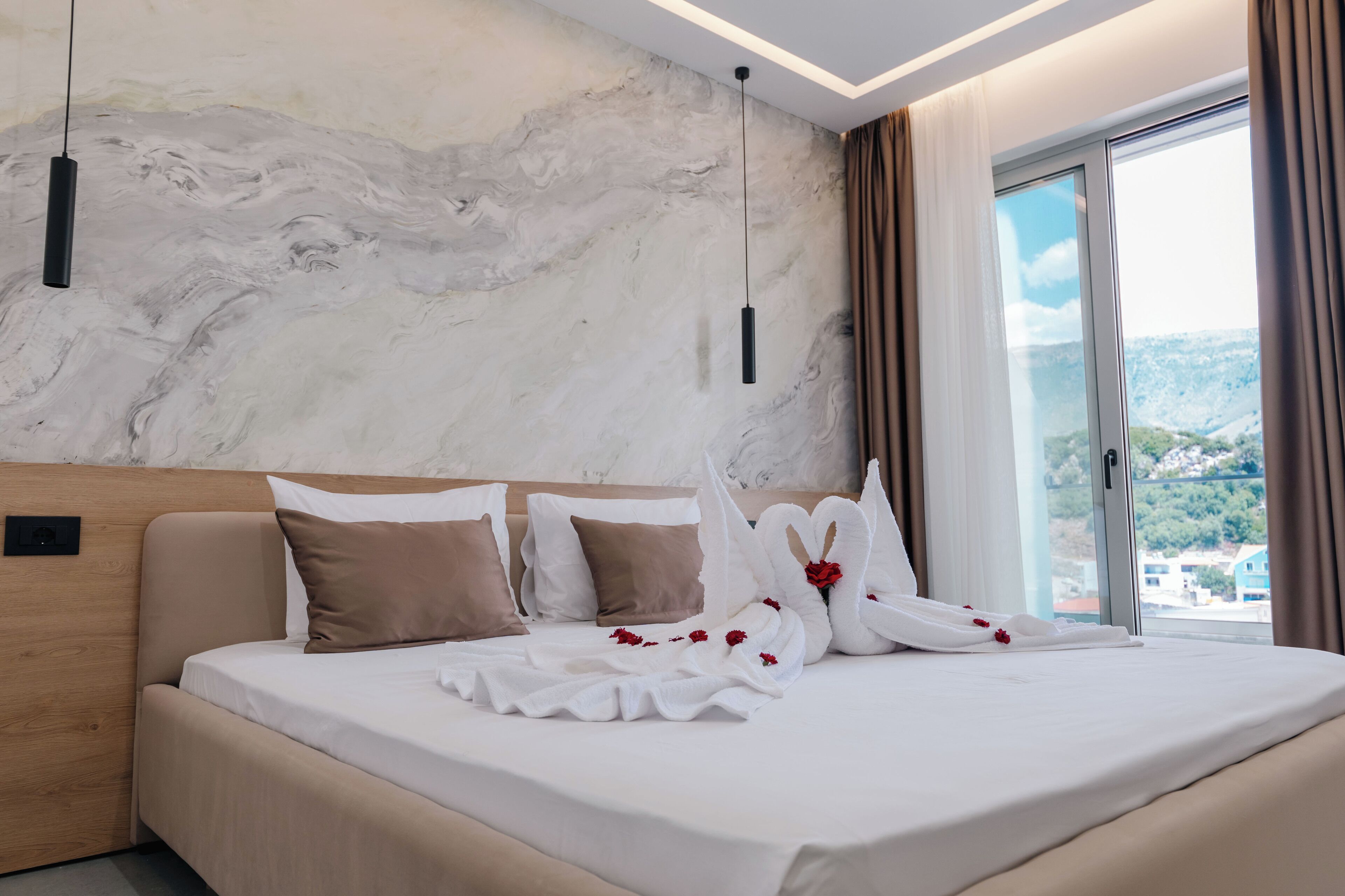 A modern hotel room with a marble wall, a large bed with white and brown bedding, and a view of a cityscape through a large window.