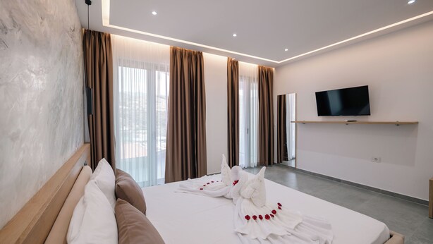 A modern hotel room with a large bed, a flat-screen TV mounted on the wall, and a view of the city through large windows. - Sun George (Himare)