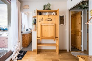 Um armário de madeira com uma placa indicando 'A Table!', em um quarto com piso de madeira, uma janela com vista para montanhas e uma porta que dá para outro cômodo.