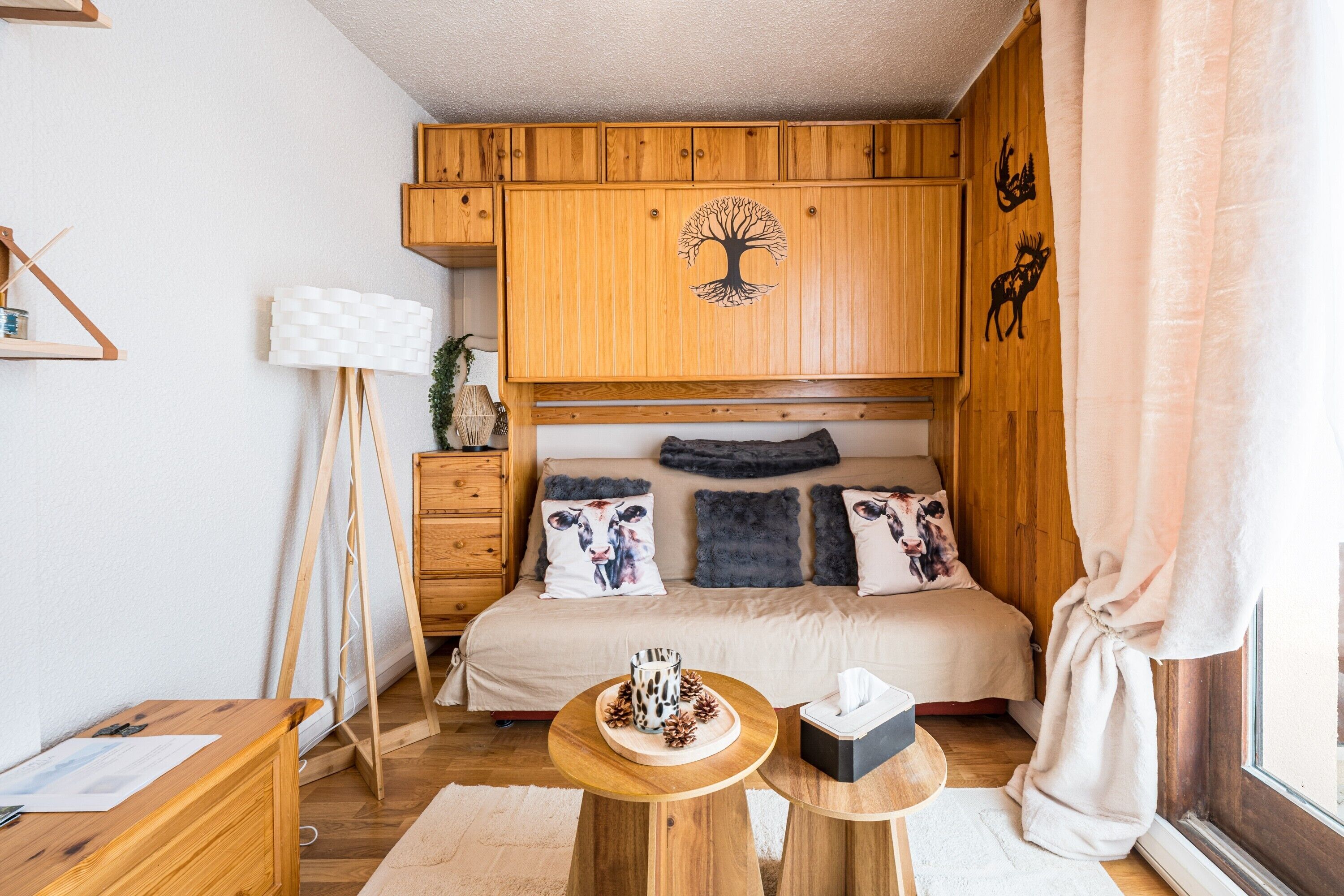 A bedroom with a wooden wardrobe, a bed with patterned pillows, a bedside table with a mug, and a window with curtains.