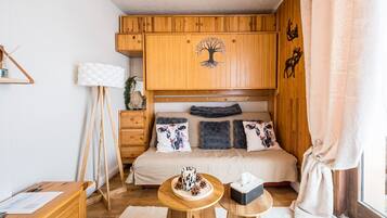 A bedroom with a wooden wardrobe, a bed with patterned pillows, a bedside table with a mug, and a window with curtains.