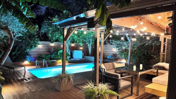 Una terraza de madera con piscina, mobiliario exterior e iluminación con luces colgantes.