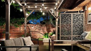Un área de estar al aire libre con una pérgola de madera, luces colgantes y un muro decorativo.