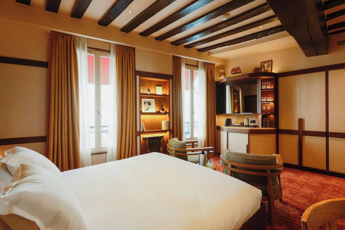 A hotel room with a large bed, wooden beams on the ceiling, a desk with a chair, and a bookshelf.