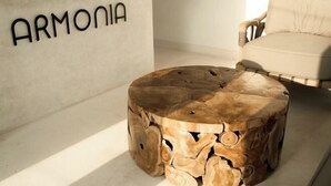 Een ronde houten salontafel met een natuurlijke houtkleur, geplaatst in een kamer met een beige bank en een aan de muur bevestigd bord waarop 'ARMONIA' staat.