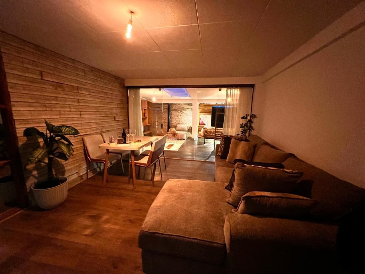 A cozy living room with a sofa, dining table, and chairs. There is a potted plant and a lamp. The room has wooden walls and a ceiling light.