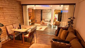 Una sala de estar moderna con piso de madera, un sofá marrón, una mesa de comedor con sillas y un área de cocina al fondo.