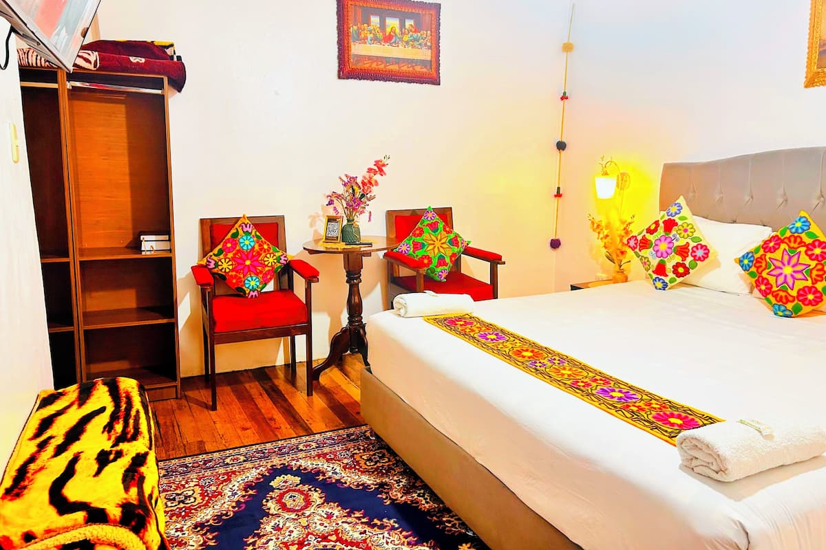 A hotel room with a bed, two chairs, a small table, a bookshelf, and a wall-mounted picture.