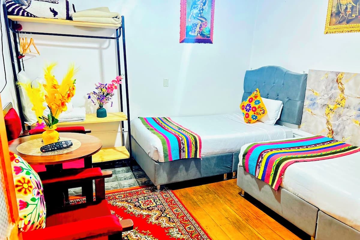 A compact room with a bed, a small table with a remote control, a chair, a shelf with folded clothes, and a decorative vase with flowers.