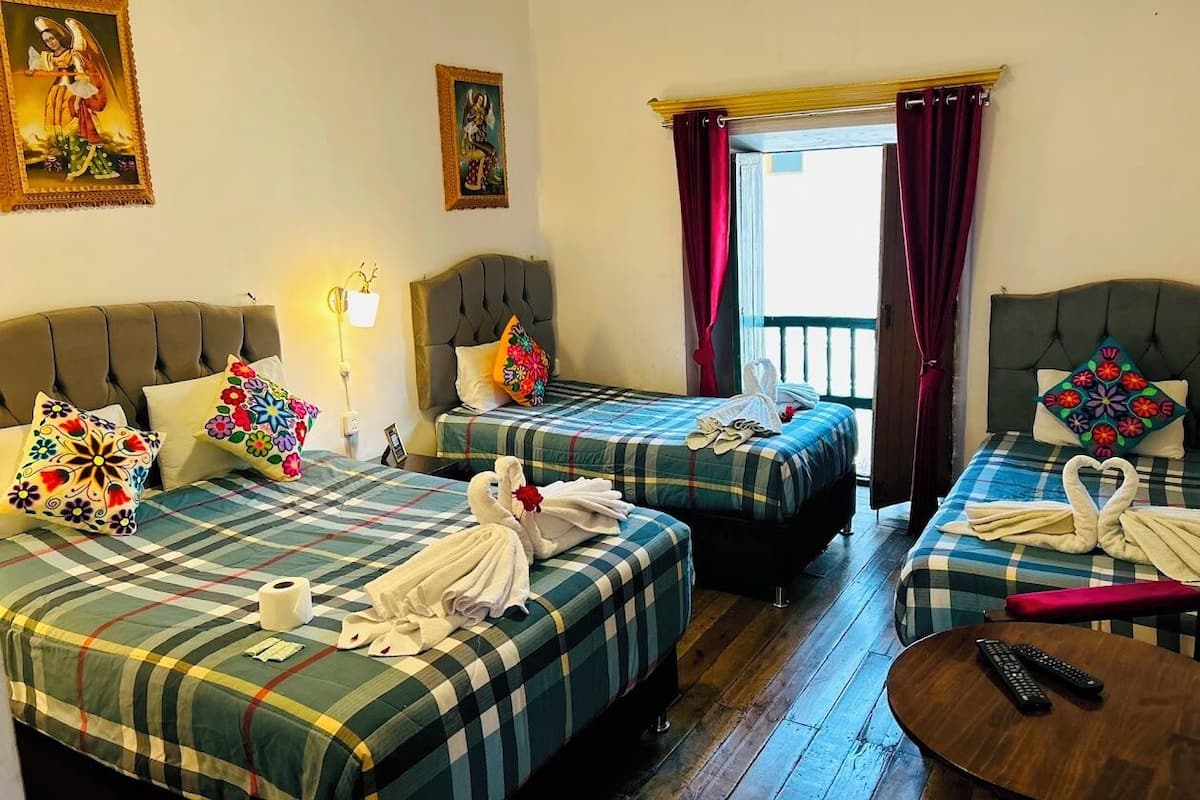 A hotel room with two beds, each with a plaid bedspread and decorative pillows. There is a small round table with a remote control, and a window with curtains.