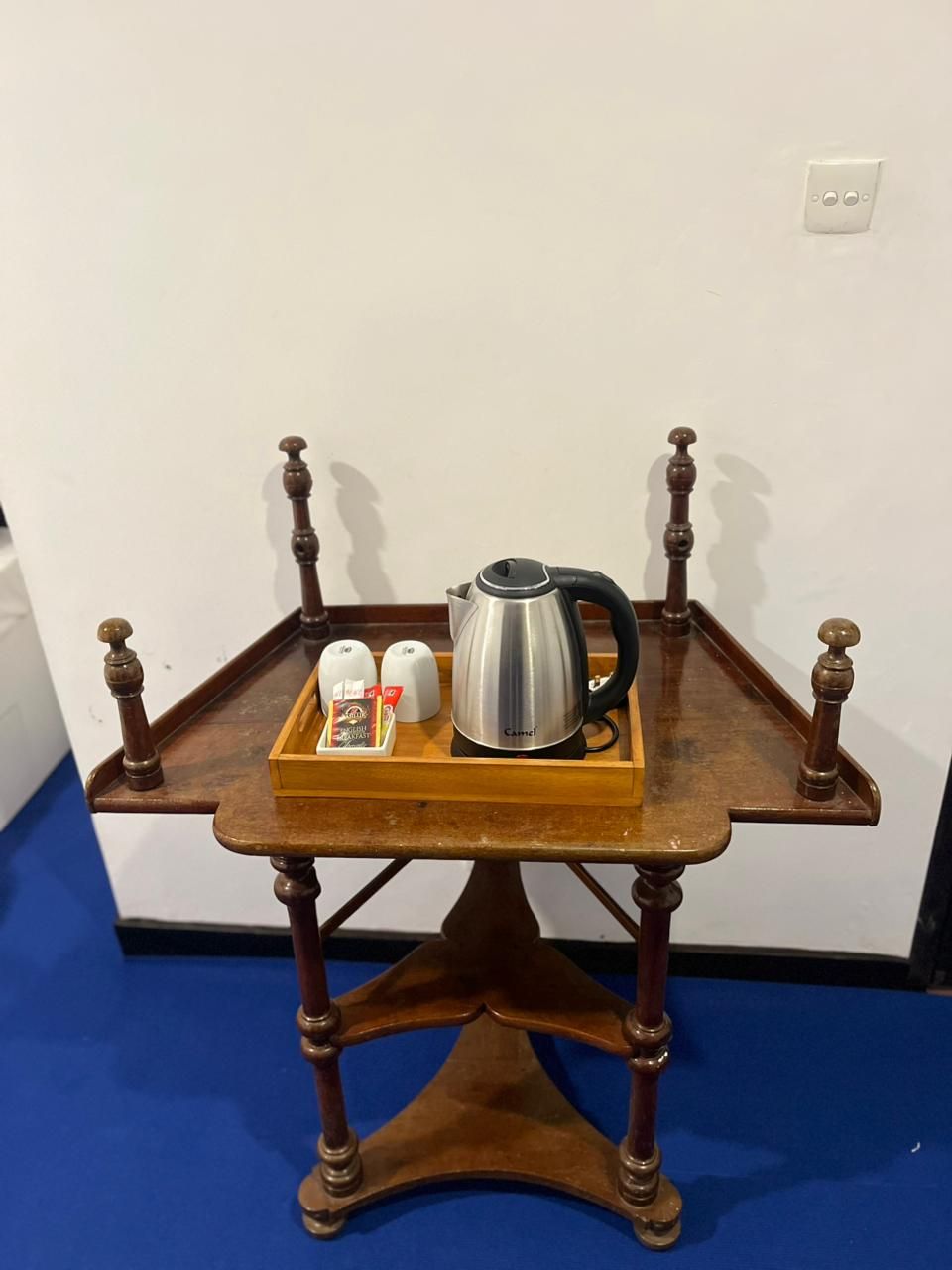 Una pequeña mesa de madera con una bandeja que contiene un hervidor eléctrico plateado, dos tazas blancas y una cajita de té.