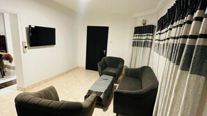 A living room with a sofa, a chair, a TV mounted on the wall, and a door. - The Crowns Lodge (Nuwara Eliya)