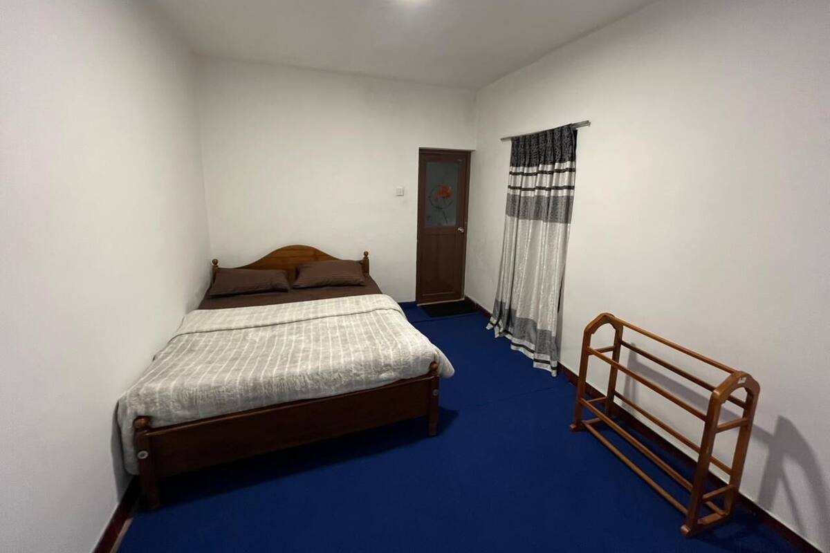 Un dormitorio con una cama de madera, una alfombra azul, una mesita de noche de madera y una ventana con cortina.