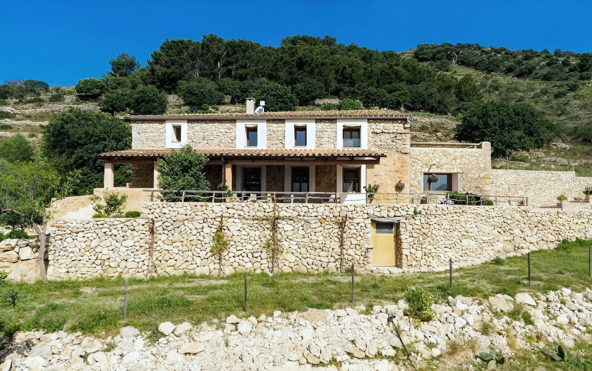 A stone house with a flat roof, multiple windows, and a stone wall surrounding the property.