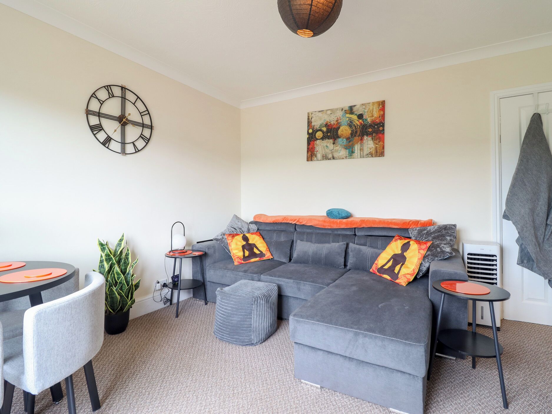 A modern living room with a grey sofa, two armchairs, a round coffee table, a small side table, a potted plant, a wall clock, and a colorful abstract painting.