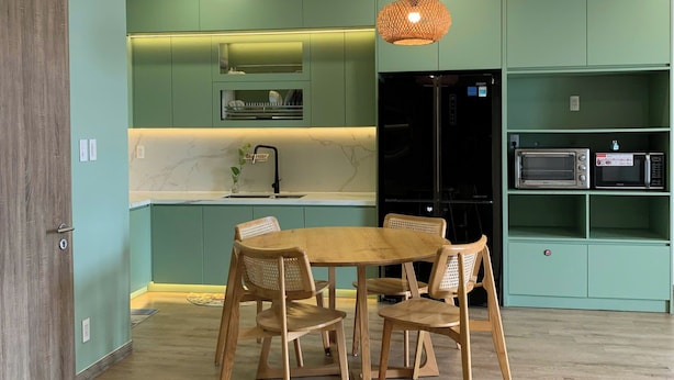 A modern kitchen with green cabinets, a wooden dining table, and a hanging light fixture. - An Corner - Sora Garden II Apartment (Ho Chi Minh City)
