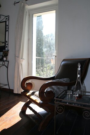 A room with a wooden rocking chair, a table with a bottle and glasses, and a view of trees through a window. - Mas de la Filoselle, a vacation home for 14 people at the foot of the Cévennes (Saint-Martin-de-Valgalgues)