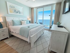 Ein Schlafzimmer mit Bett, Nachttischen, einem Schminktisch, einem Kleiderschrank, einem Balkon mit Meerblick und einem Deckenventilator.