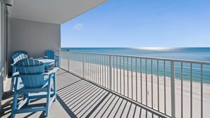 A balcony with blue Adirondack chairs and a table, overlooking a beach with a railing.