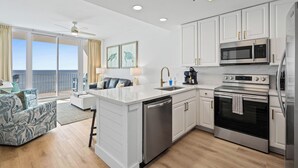 A modern kitchen with white cabinets, stainless steel appliances, and a large island with a dishwasher. There is a balcony with a view of the ocean.