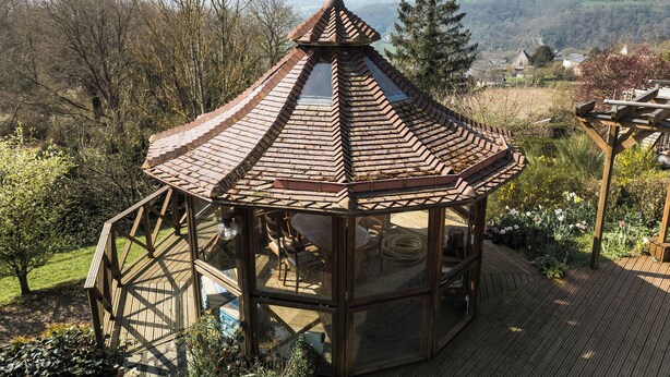 Ein hölzerner Gartenpavillon mit einem Ziegeldach, umgeben von einem Garten mit verschiedenen Pflanzen und Blumen.